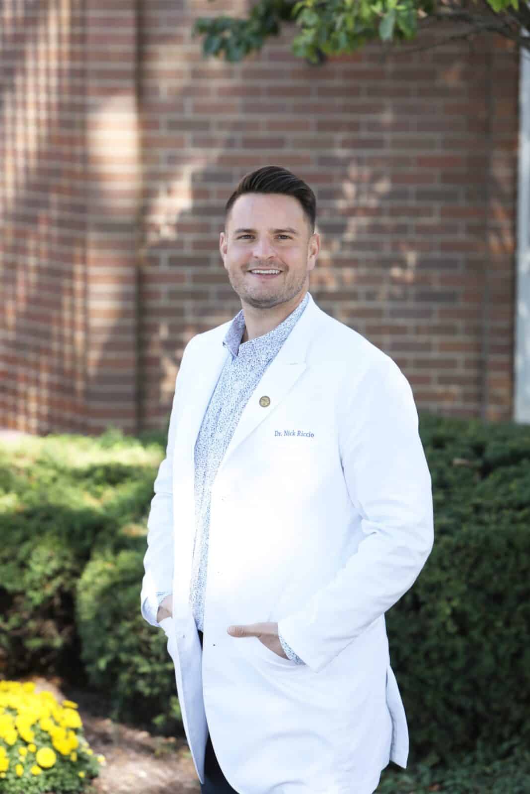 Dr. Nick Riccio, orthodontist in Brookfield, wearing a white coat, smiling outdoors near yellow flowers, representing personalized orthodontic care at Hinsdale Orthodontics.
