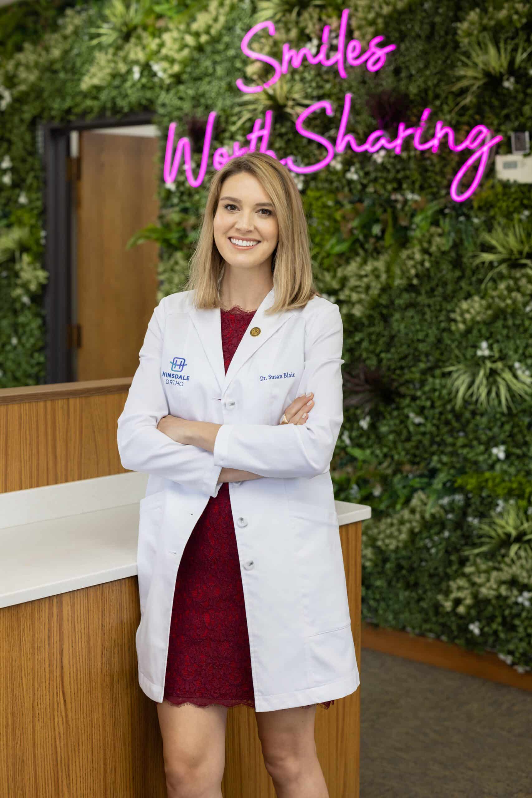 Dr. Susan Blair, orthodontist at Hinsdale Orthodontics, smiling in a white coat against a green wall with "Smiles Worth Sharing" sign, representing Brookfield orthodontic care and personalized treatments.